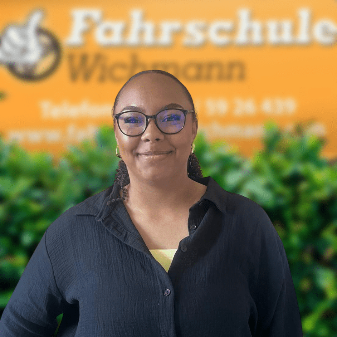 Charline, Fahrlehrerin der Fahrschule Wichmann in Oyten, begleitet dich kompetent beim Autoführerschein, Motorradführerschein, LKW- und Anhängerführerschein sowie der Fahrlehrerausbildung – auch in Bassen und Borstel oder per Intensivfahrschule. - Wichmann Fahrschule Oyten - Autoführerschein LKW Führerschein Motorradführerschein Charline, Fahrlehrerin der Fahrschule Wichmann in Oyten, steht vor dem gelben Firmenschild mit Logo und grüner Hecke.