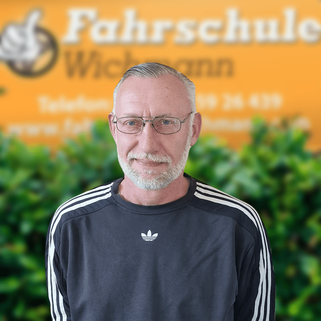Peter, Fahrlehrer der Fahrschule Wichmann in Oyten, begleitet Schüler aus Oyten, Bassen und Borstel beim Autoführerschein, Motorradführerschein, LKW- und Anhängerführerschein sowie in der Intensivfahrschule und Fahrlehrerausbildung. - Wichmann Fahrschule Oyten - Autoführerschein LKW Führerschein Motorradführerschein Peter, Fahrlehrer der Fahrschule Wichmann in Oyten, steht vor dem Logo der Fahrschule mit grünem Busch im Hintergrund.