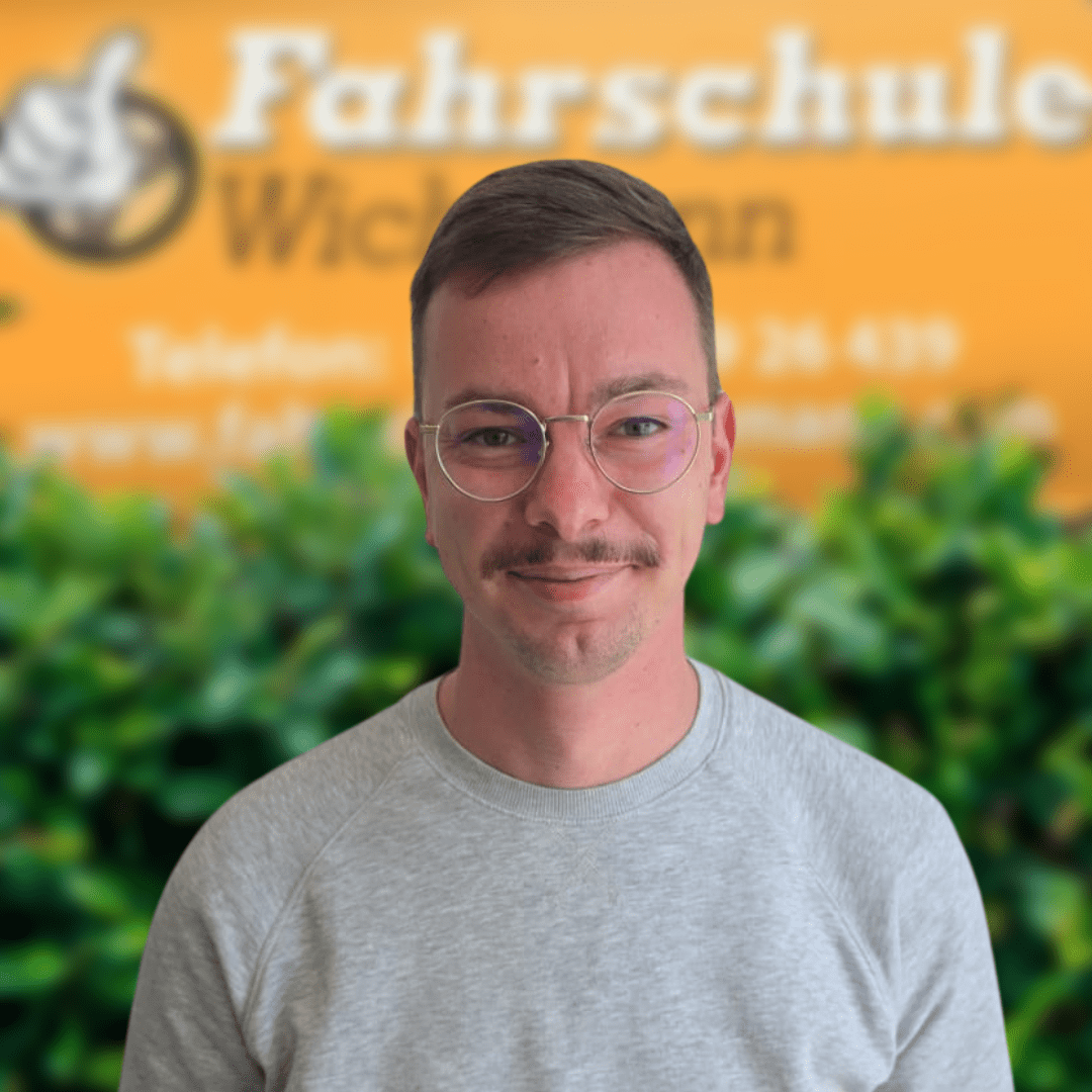 Fahrlehrer Sofiane der Fahrschule Wichmann Delmenhorst vor grünem Busch und gelbem Firmenschild.
