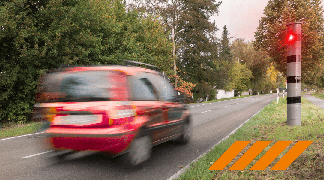 Blurred red car speeds along a tree-lined road beside a railroad crossing signal with orange hazard stripes on the verge.
